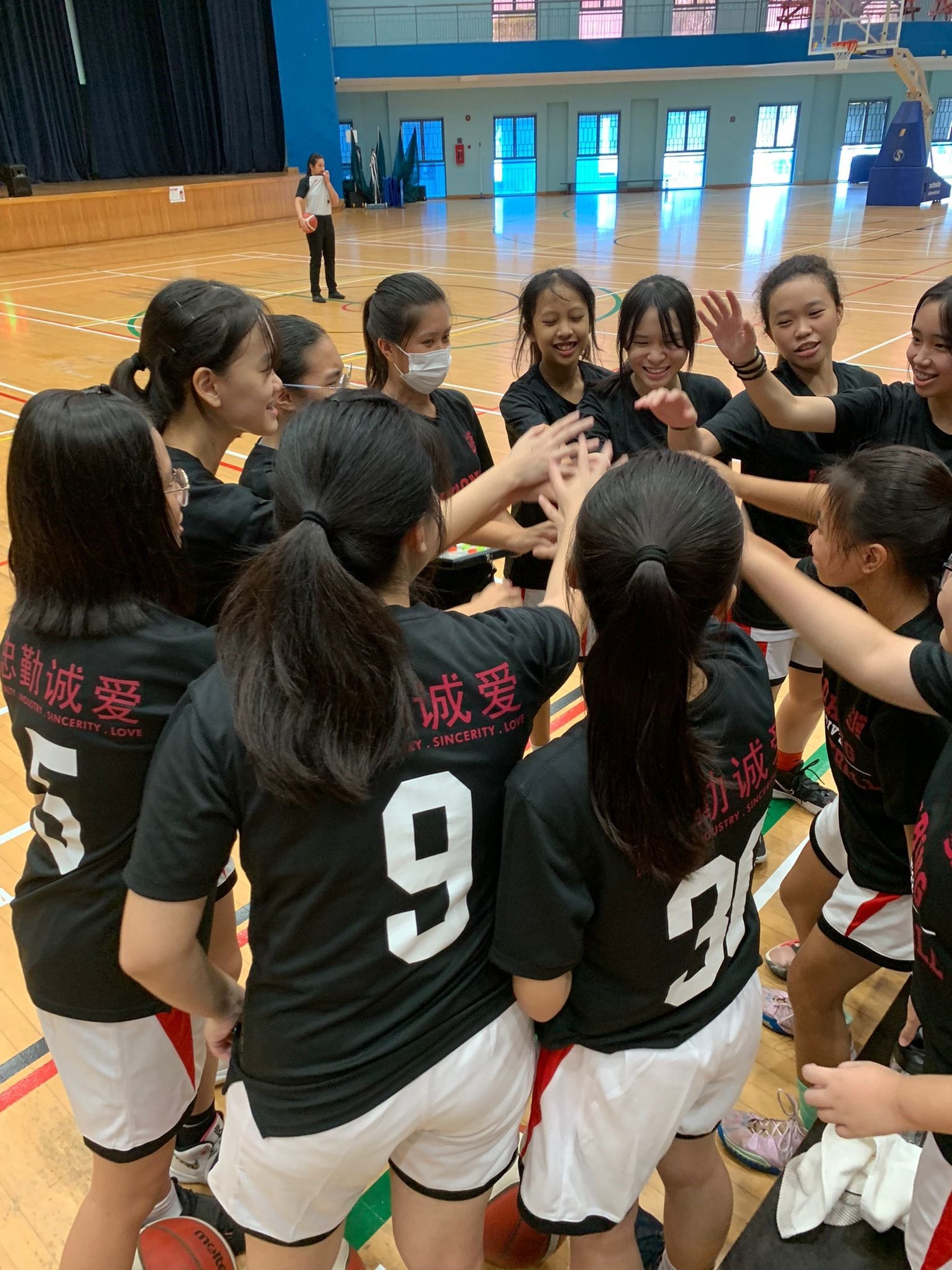 A girls basketball team in black uniforms huddles together with hands raised in the center of an indoor court.